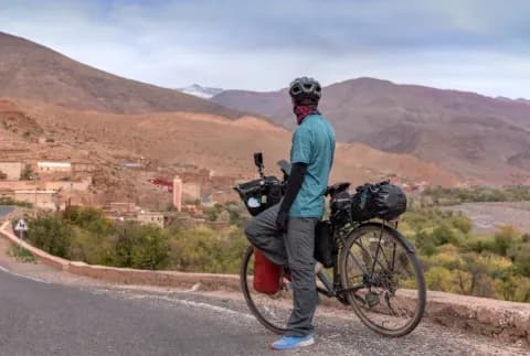 Ein Fahrradfahrer steht am Straßenrand mit seinem Fahrrad und blickt auf ein Dorf, braune Bergen und grüne Wälder.
