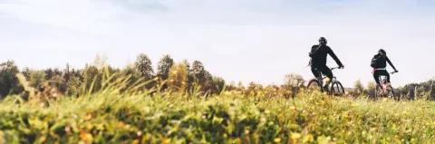 Zwei Radfahrer radeln über eine grüne Wiese bei bewölktem Himmel