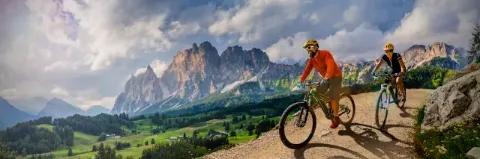 Ein Paar radelt auf E-Bikes auf einer Schotterpiste vor grüner Wiese und Bergkulisse bei bewölktem Himmel