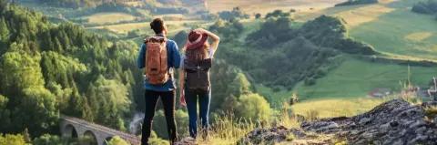 Pärchen mit Wanderrucksack blickt in die Ferne über eine grüne Hügellandschaft