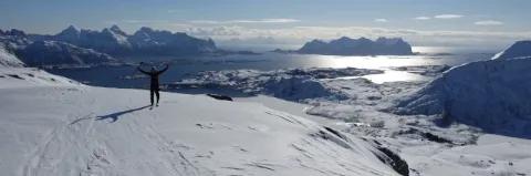 Skifahrer jubelt auf Skipiste und blickt auf den schneebedeckten Fjord unter blauem Himmel und Sonnenschein