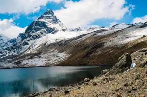Blick über einen blauen Gebirgssee zum spitzen und felsigen Gipfel des Alpamayo, der an vielen Stellen noch mit etwas Schnee bedeckt ist.
