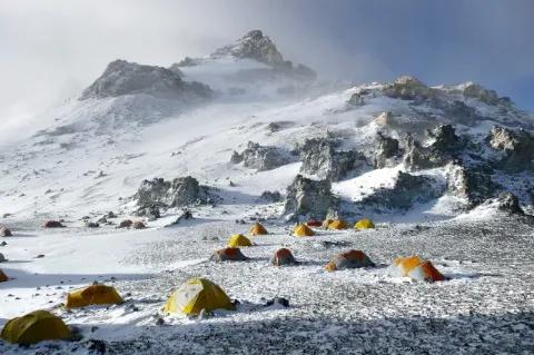 Eine Vielzahl kleiner, gelber Zelte steht auf einer von Schnee bedeckten Ebene vor einem in den Himmel aufragenden Berg mit mehreren felsigen Spitzen.