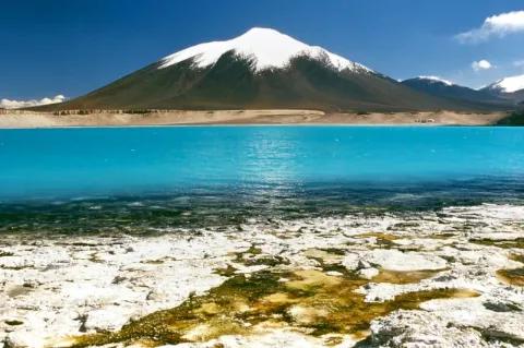 Blick über türkisblaue, glatte Seeoberfläche zu einem in den Himmel aufragenden Vulkan. Der oberen Teil des Vulkans ist mit Schnee bedeckt.