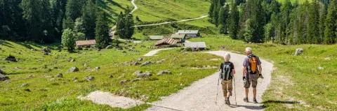 Paar wandert auf einem Forstweg hinab in Richtung Berghütte neben grünen Wiesen und Wäldern