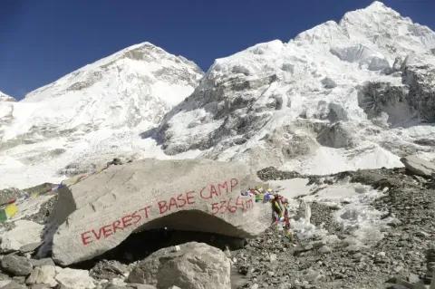 Auf einem großen Fels steht mit roter Schrift "Everst Base Camp". Der Fels liegt auf einer steinigen Ebene. im Hintergrund sind schneebedeckte Gipfel zu sehen.