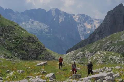 Drei Wanderer steigen über einen grünen Hang in einem Gebirge auf.