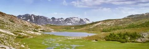 Landschaft mit grüner Wiese, grünem Berghang, See und Bergkette mit wenig Schnee im Hintergrund