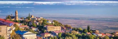 Stadt mit Häusern und Kirche auf einem Hügel mit Bergkette im Hintergrund unter bewölktem Himmel