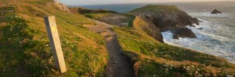 Wegweiser an einem Küstenpfad mit grüner Wiese und tosender Brandung bei Sonnenuntergang