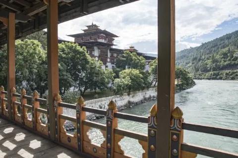 Blick von einer Brücke aus über einen Fluss auf ein buddhistisches Kloster. Vor dem Dzong wachsen grüne Bäume.
