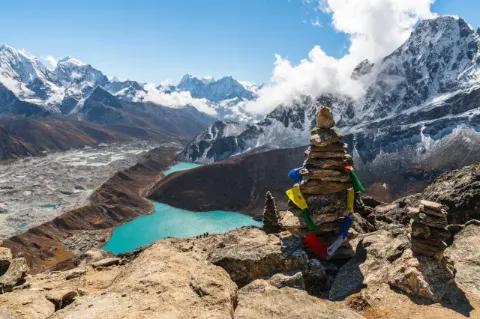 Im Vordergrund steht ein von Gebetsfahnen geschmückter Steinturm auf einem Felsvorsprung. Dahinter ist das beeindruckende Bergpanorama des Gokyo zu sehen. Weiter unten liegt ein türkisblauer Gebirgssee.