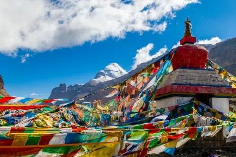 Blick auf einen schneebedeckten, spitzen Berggipfel im Hintergrund. Im Vordergrund sind Teile eines buddhistischen Tempels zu sehen, der mit vielen bunten Gebetsflaggen geschmückt ist.