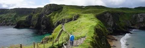 Wanderer läuft auf einem abgesperrten Küstenwanderweg zwischen grüner Wiese und blauem Meer