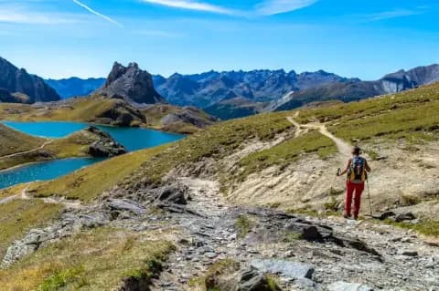 Wanderer mit Rucksack und Wanderstöcken läuft über eine grüne, felsige Hochebene auf einen blauen See zu