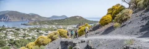 Vier Wanderer beim Aufstieg auf den Stromboli. Das Meer und mehrere grün bewachsene Hügel liegen im Hintergrund.