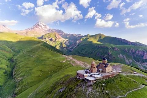 Auf einem grünen Berg ist eine Kirche mit Kuppel und Glockenturm. Im Hintergrund sind hohe Berge.