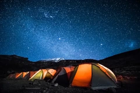Eine Reihe leuchtend oranger Zelte wird in einer dunklen Nacht angeleuchtet. Am tief dunkelblauen Himmel ist die Milchstraße zu sehen.