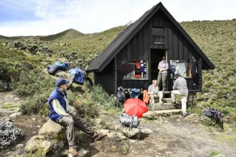 Eine Gruppe Wanderer sitzt vor einer einfachen Holzhütte an einem Berg. Die Landschaft ist niedrig grün bewachsen und felsig.
