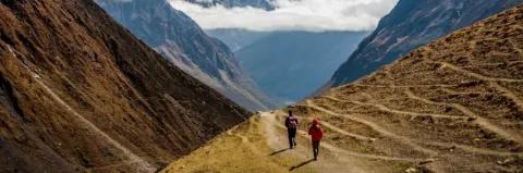 Zwei Wanderer auf Wanderweg, der durch ein Tal führt, mit schneebedeckten Bergen im Hintergrund