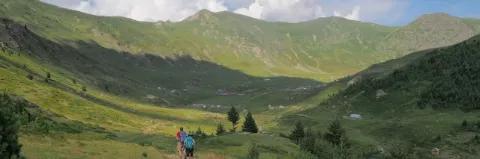 In einem grünen Tal umgeben von grün bewachsenen Bergen wandern zwei Wanderer.