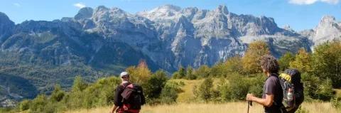 Zwei Wanderer gehen auf einer grasigen Wiese. Im Hintergrund sind Berge aus grauem Gestein.
