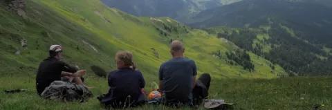 Drei Wanderer sitzen auf einer Wiese am Berg. In der Ferne geht es runter in ein grünes Tal.