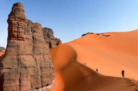 Vier Wanderer gehen auf dem Kamm einer Sanddüne. Links sind bizarre rötliche Felsformationen.