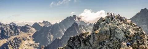 Felsiger Gipfel mit Menschen vor einer felsigen Bergkulisse unter trübem blauen Himmel mit Wolken