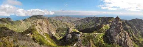 Ein zerklüftetes Bergpanorama. Die Berge sind grün bewachsen. In der Ferne ist das Meer.