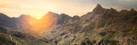 Die Sonne geht hinter einer Bergkette unter. Die Berge sind teils grün bewachsen.