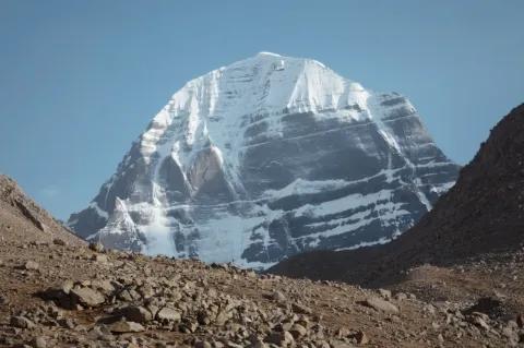 In der Mitte ist der hohe, schneebedeckte Gipfel des Kailash. Rechts und links sind steinige Berge.