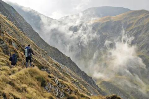 Zwei Wanderer mit großen Rucksäcken gehen auf einem schmalen Pfad entlang eines steilen Hangs in einem mit Gras bewachsenen Gebirge. Zwischen den Bergen wabert Nebel.