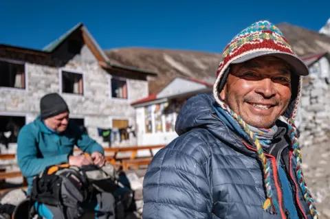 Der nepalesische Reiseleiter Harka Tamang blickt lächelnd in die Kamera. Er hat eine blaue Daunenjacke und eine Mütze mit bunten Mustern an und steht vor einem grauen Gebäude.