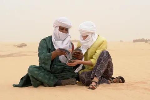 Zwei Menschen mit Kopftüchern sitzen im Sand und schauen sich gemeinsam ein Magazin an.