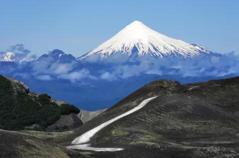 Eine schmale Straße führt über einen Hügel. Im Hintergrund steht ein schneebedeckter Vulkan