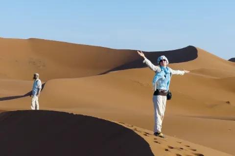Eine Frau steht auf einer Düne und blickt in Richtung Kamera. Sie breitet die Arme aus und lacht.