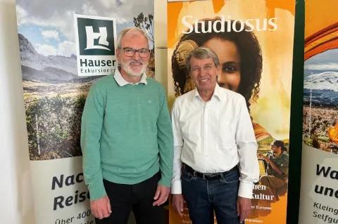 Zwei Männer stehen vor zwei Werbeflächen der Firmen Studiosus und Hauser Exkursionen. Beide schauen in die Kamera und lächeln.