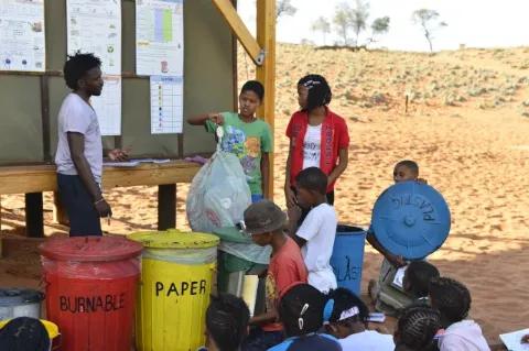 Ein Gruppe Kinder steht um mehrere bunte Abfalleimer herum. Ein Kind hält eine Mülltüte in der Hand.