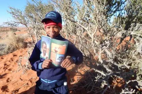 Ein Junge hat eine Zeitschrift in den Händen, die er in die Kamera zeigt. Im Hintergrund ist ein Baum und Wüstenlandschaft zu sehen.