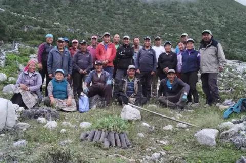 Eine große Gruppe Menschen steht auf einer grünen Wiese. Vor der Gruppe liegen kleine Baumsetzlinge.
