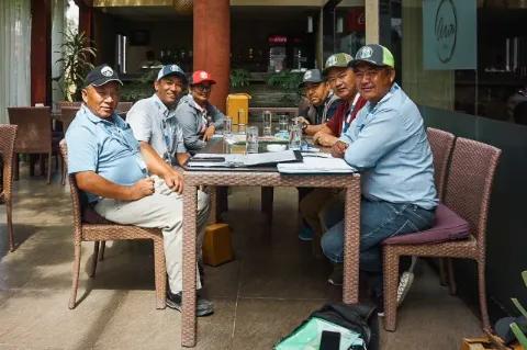 Sechs Männer mit Mützen sitzen auf Stühlen an einem Tisch und schauen in die Kamera.