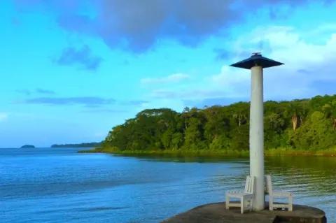 Ein kleiner grauer Leuchtturm steht auf einem Plateau auf dem Wasser. Im Hintergrund ist ein Teil einer grünen Insel zu sehen.