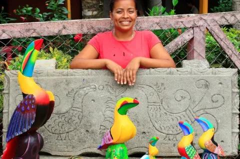 Eine Frau steht hinter einer steinernen Platte und lacht in die Kamera. Vor der Platte sind verschiedene angemalte Holzfiguren aufgestellt.