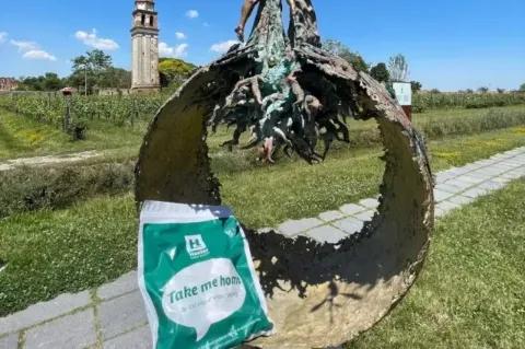 Der grün bedruckte Take me home-Beutel liegt auf einer Skulptur. Im Hintergrund sind eine Steinkirche und Weinfelder zu sehen.