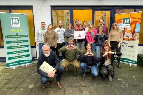Das Hauser-Team steht als Gruppe vor dem Bürogebäude und schauen in die Kamera. Ein paar der Mitarbeitenden formen Herzen mit ihren Händen.