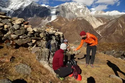 Ein Mann mit orangener Jacke beugt sich zu zwei sitzenden Männern und teilt sein Essen. Die Landschaft ist felsig mit schneebedeckten Gipfeln im Hintergrund