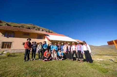 Eine große Gruppe steht auf grünem Rasen vor einem einfachen Haus und schaut in die Kamera.