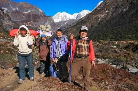Eine Gruppe aus vier Männern die teilweise Gepäck auf dem Rücken tragen steht auf felsigem Untergrund und blickt in die Kamera. Im Hintergrund sind schneebedeckte Gipfel zu sehen.