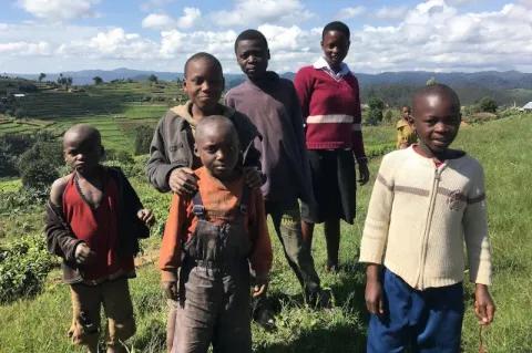 Sechs Kinder stehen auf einer Wiese und blicken in die Kamera. Im Hintergrund ist eine grüne Hügellandschaft zu sehen.
